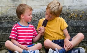 two boys sharing an ice cream next to a wall